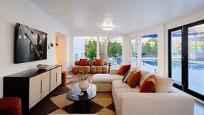 A modern living room with a sectional sofa, a flat-screen TV, a coffee table, and a view of a swimming pool. - Sunburst Estate - Pool - Hot Tub - Tons of Games! (Scottsdale)