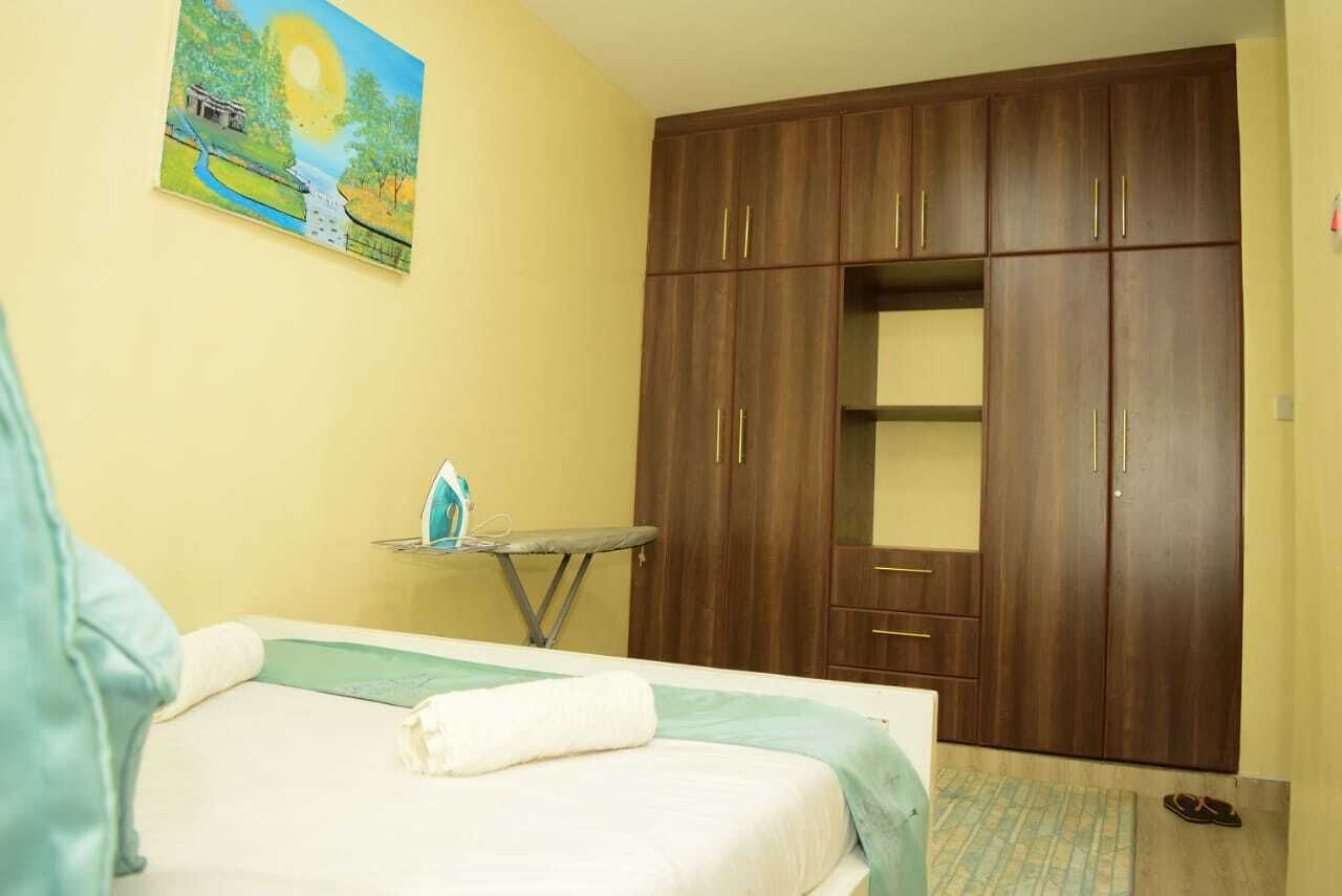 A bedroom with a bed, a wooden wardrobe, a small table with an ironing board, and a painting on the wall.