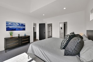 A modern bedroom with a large bed, a dark dresser, a wall-mounted TV, and a hallway leading to a bathroom.