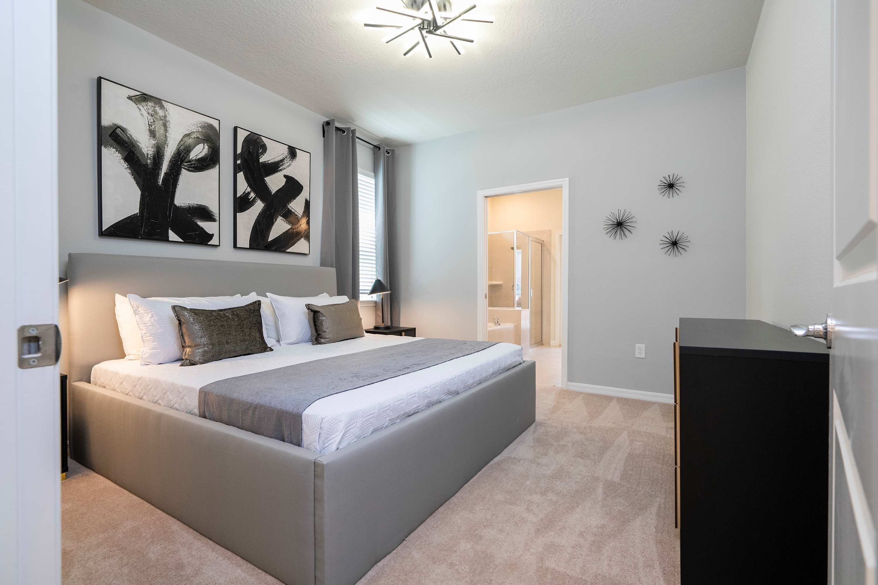 A bedroom with a large bed, a nightstand, a dresser, abstract wall art, and a ceiling light fixture.