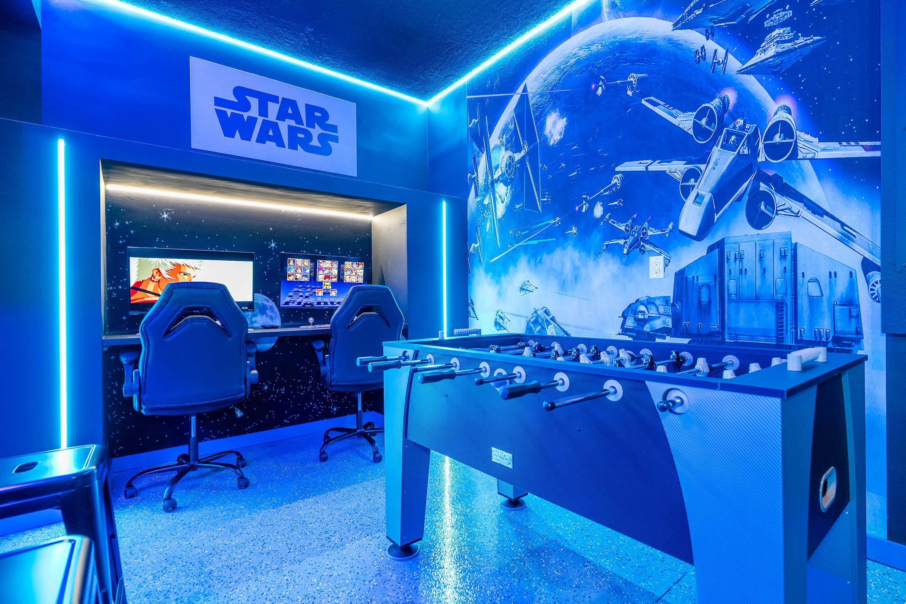 Een kamer met een tafelvoetbaltafel, twee gaming stoelen, een muur met Star Wars thema en een groot raam met uitzicht op de lucht.