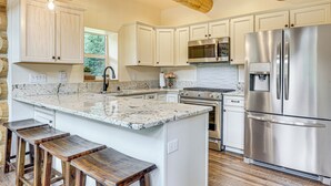 A modern kitchen with white cabinetry, a large island with bar stools, stainless steel appliances, and exposed wooden beams. - Log Cabin w/ Wraparound Deck in Breckenridge! (Breckenridge)