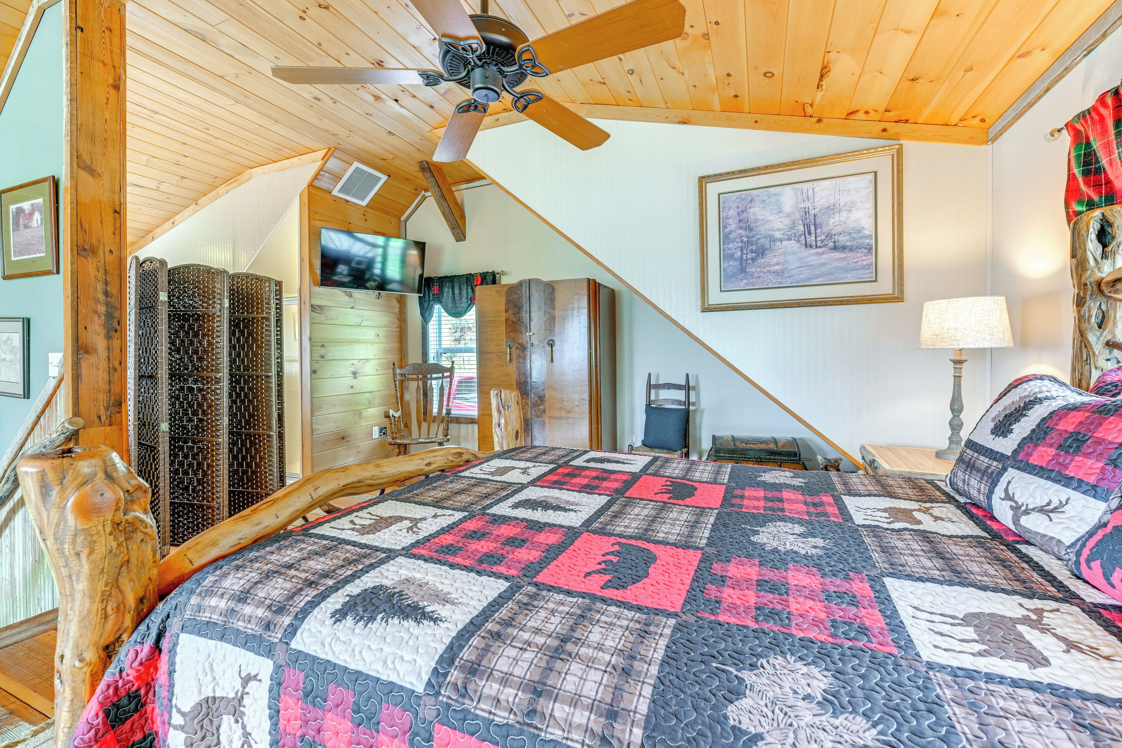 A cozy bedroom with a wooden ceiling, a quilted bedspread, a ceiling fan, and a painting on the wall.