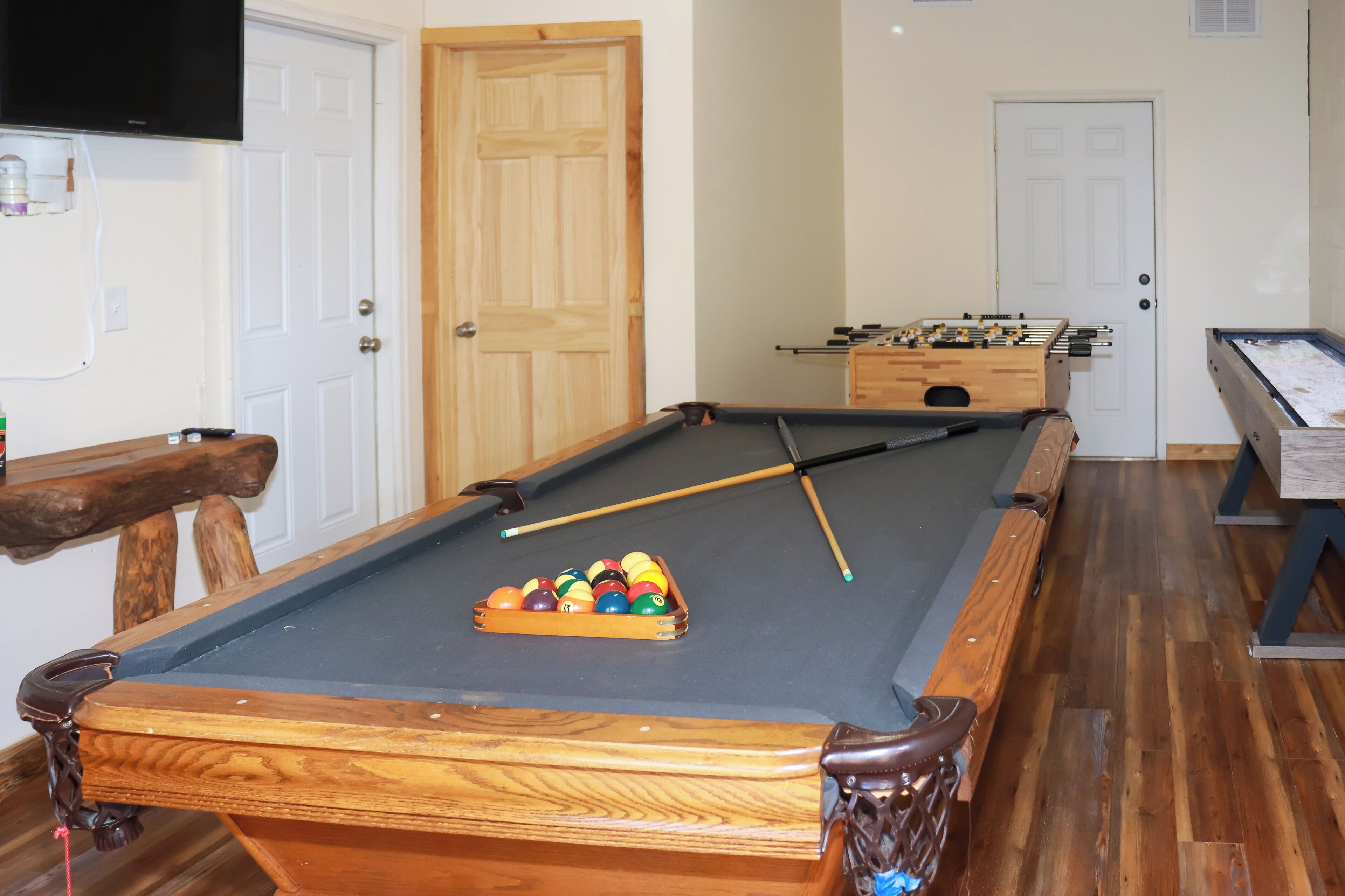 A pool table with balls, a foosball table, and a billiards table in a room with wooden flooring and white walls.