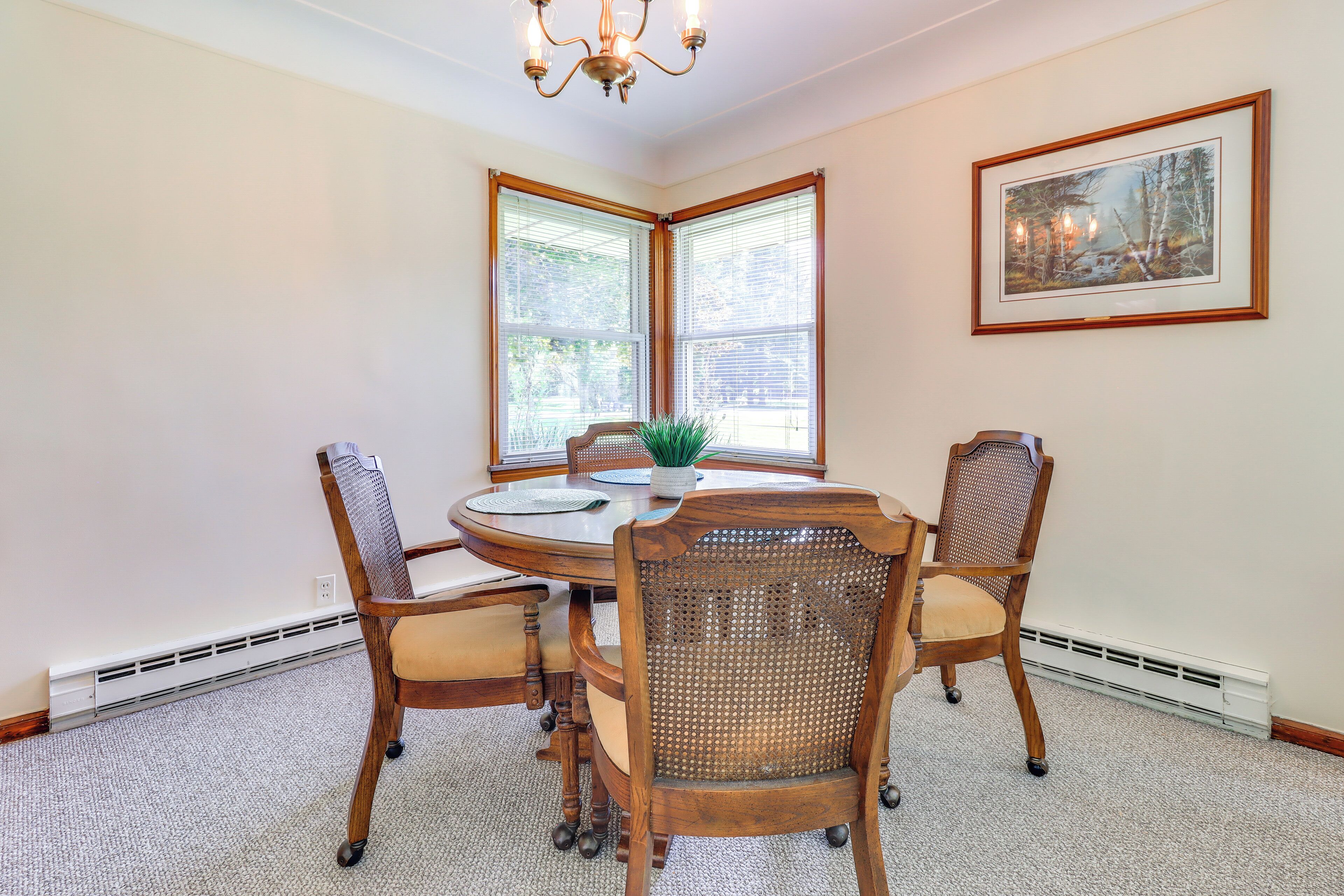 A dining area with a round table, four wooden chairs, a chandelier, and a framed picture on the wall.