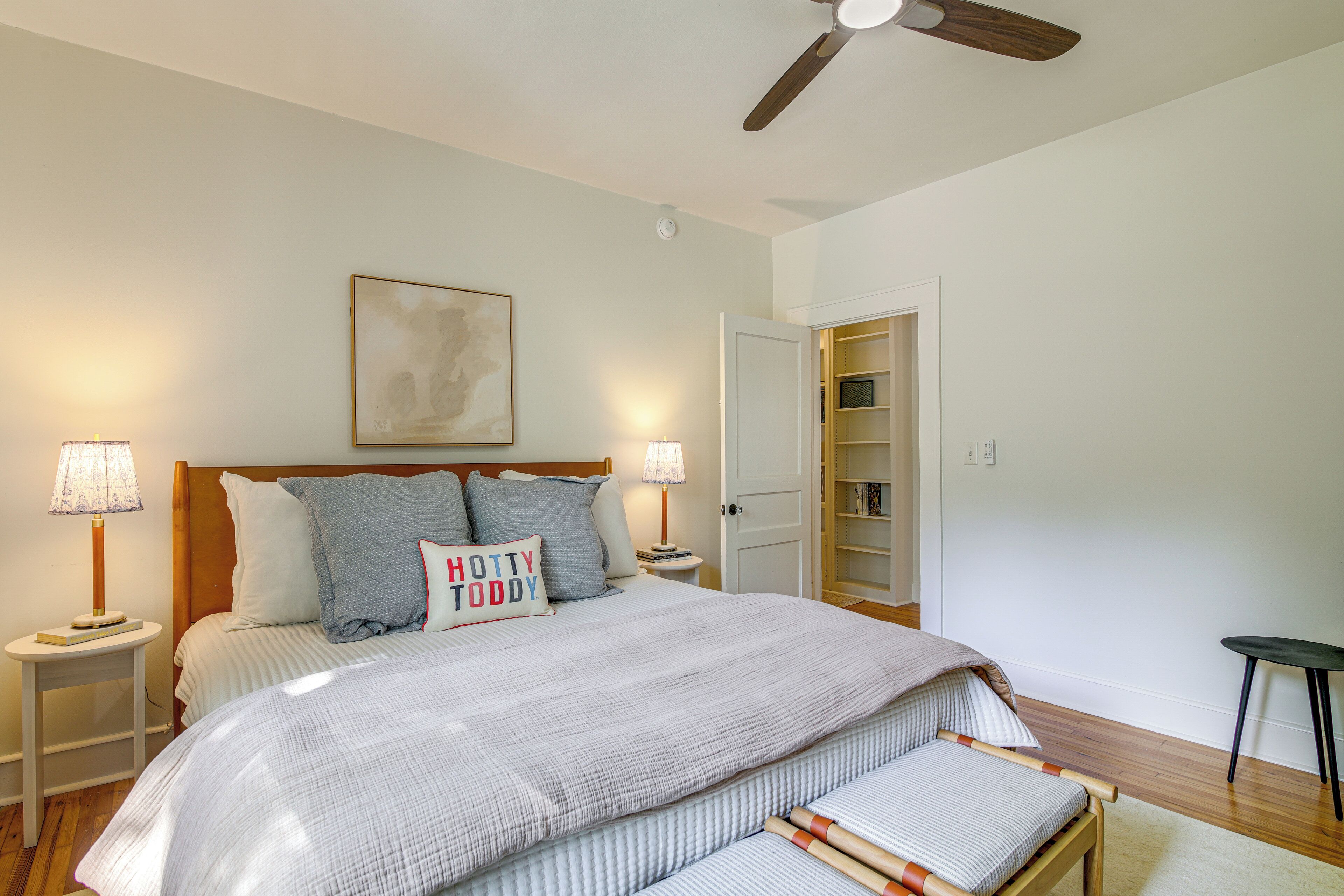 A bedroom with a bed, bedside lamps, a ceiling fan, a nightstand, a bedside table, a bench, and a door leading to a closet.