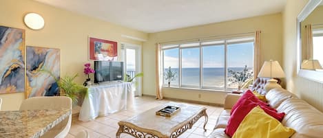 A living room with a marble table, a sofa with colorful cushions, a TV, and a large window with a view of the sea.