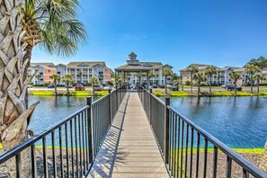 A wooden walkway leading to a gazebo over a body of water, surrounded by multi-story buildings with balconies and palm trees. - Screened Porch & Community Perks: Myrtle Beach Gem (Myrtle Beach)