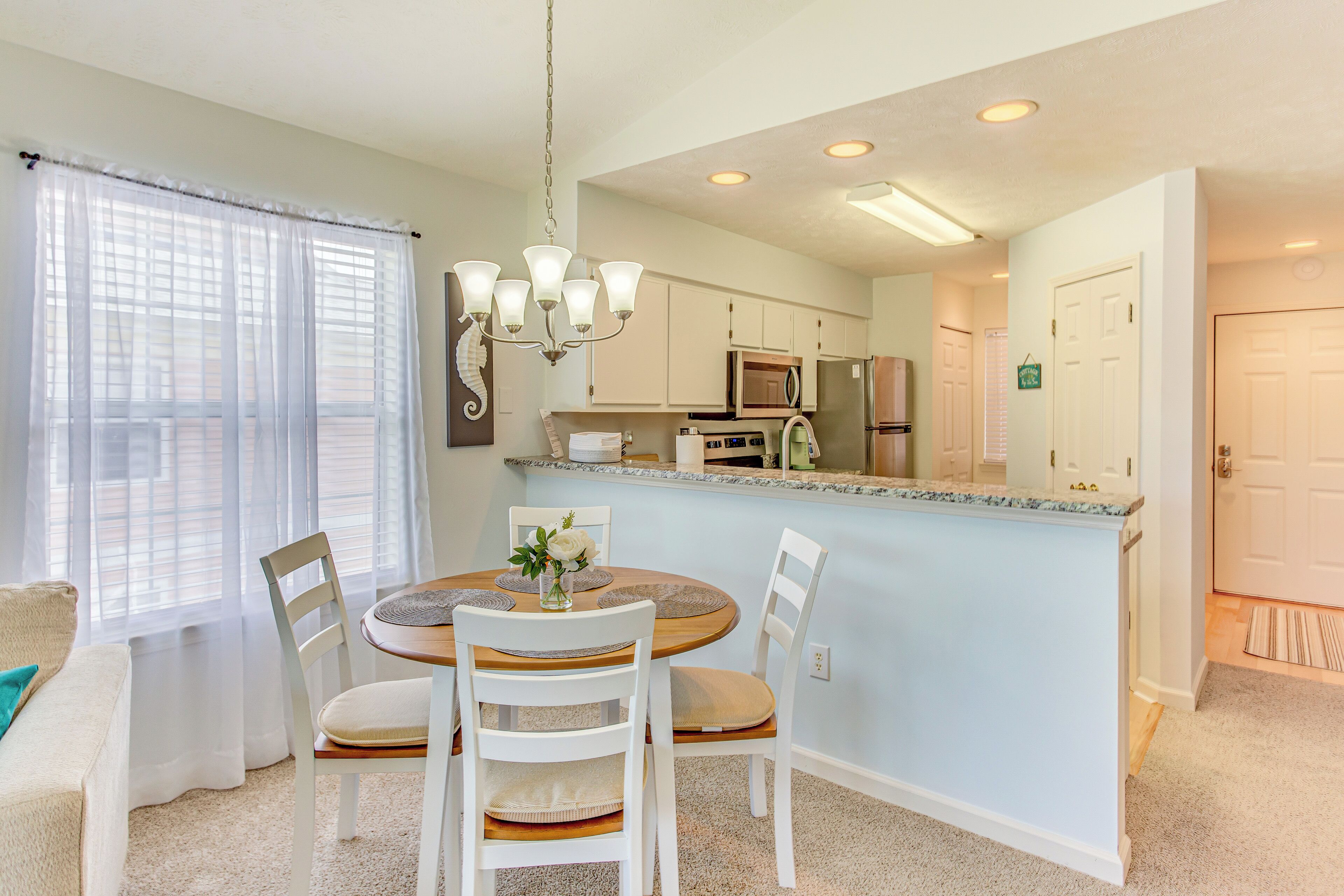 A dining area with a round table, white chairs, a chandelier, and a kitchen in the background.