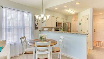 A dining area with a round table, white chairs, a chandelier, and a kitchen in the background.