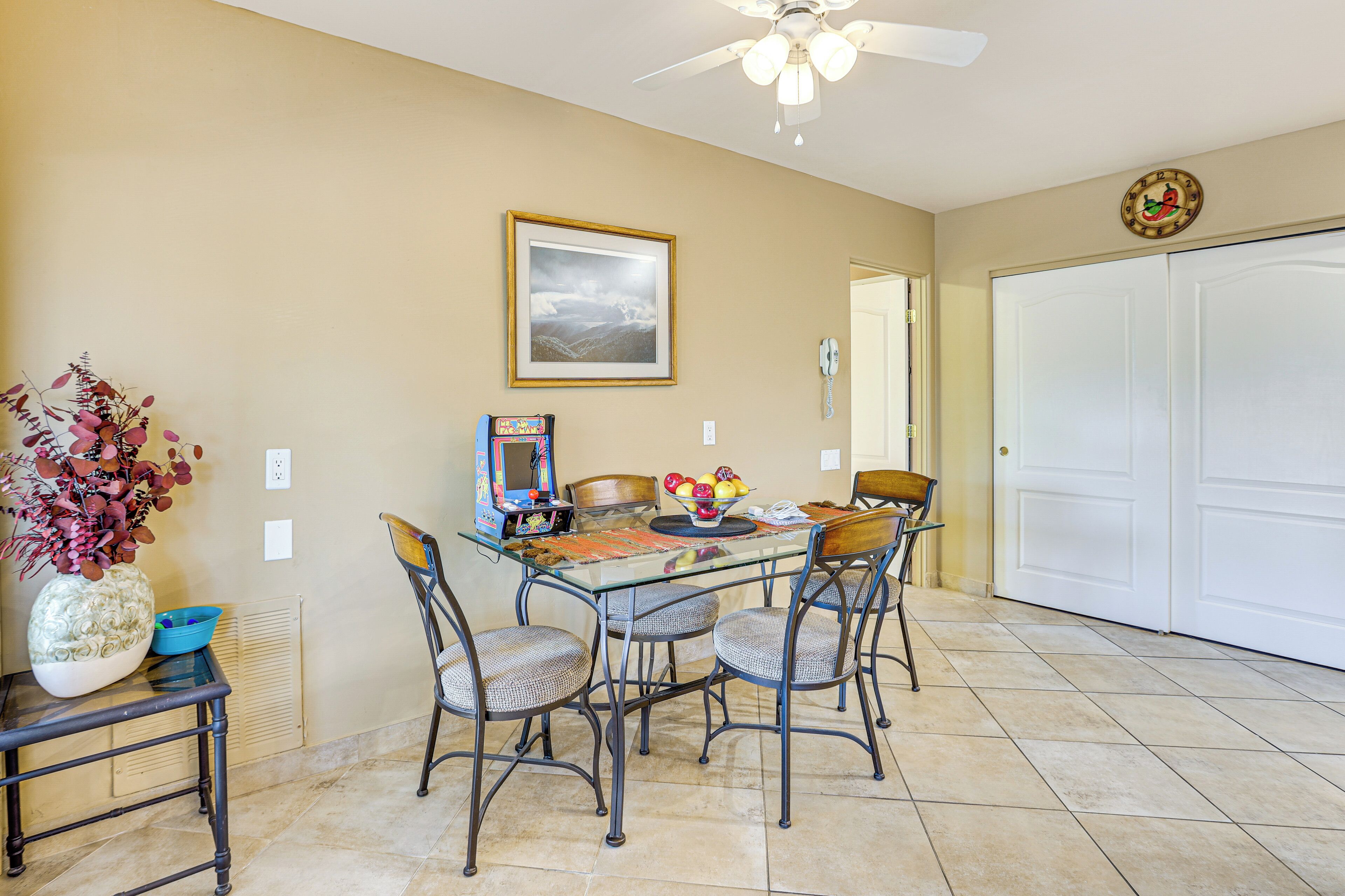 A dining area with a glass table, four chairs, a vase with flowers, a framed picture, and a ceiling fan.