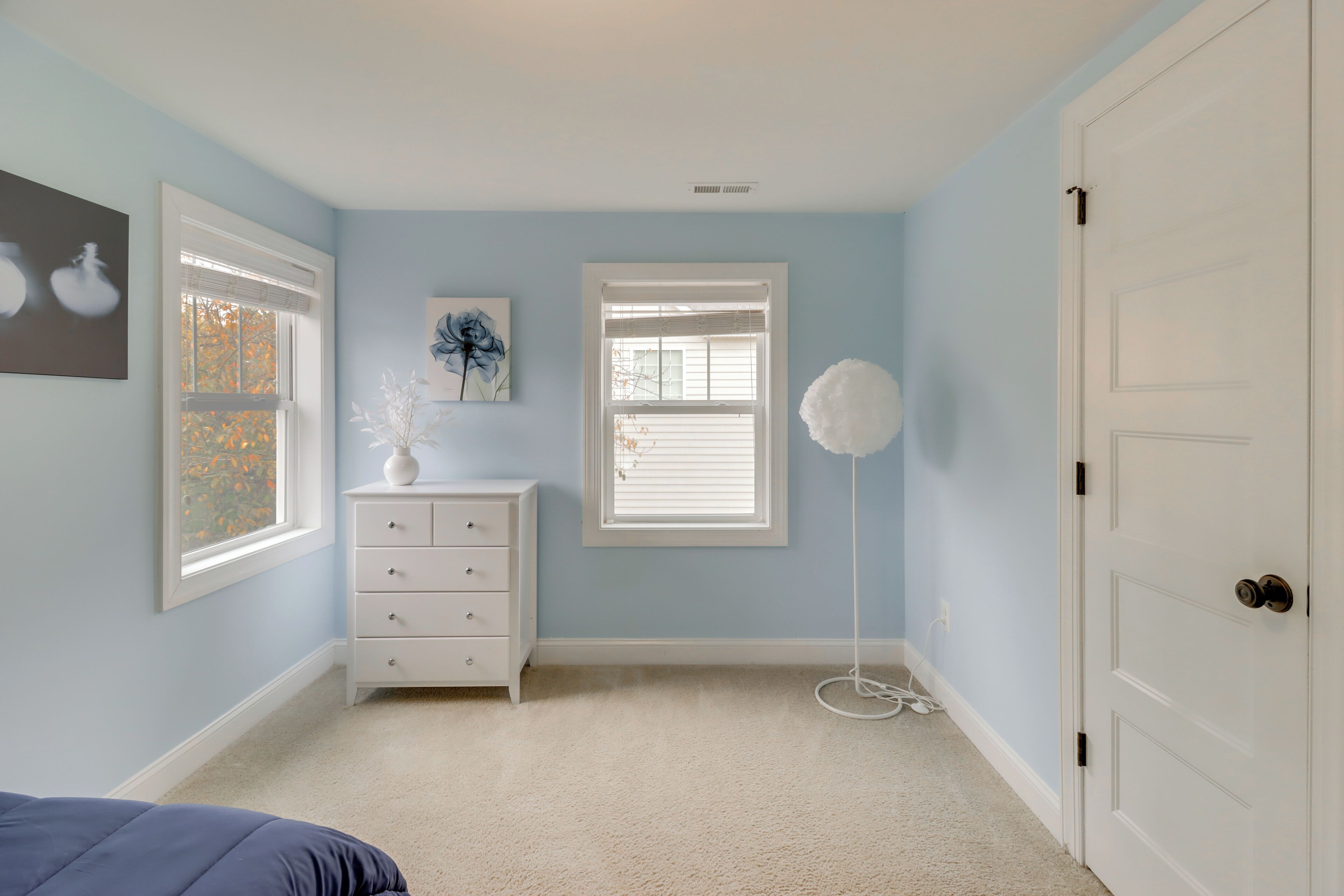 Un dormitorio con paredes azul claro, un aparador blanco, dos ventanas con persianas, una puerta blanca y una cama con cubrecolcha azul.