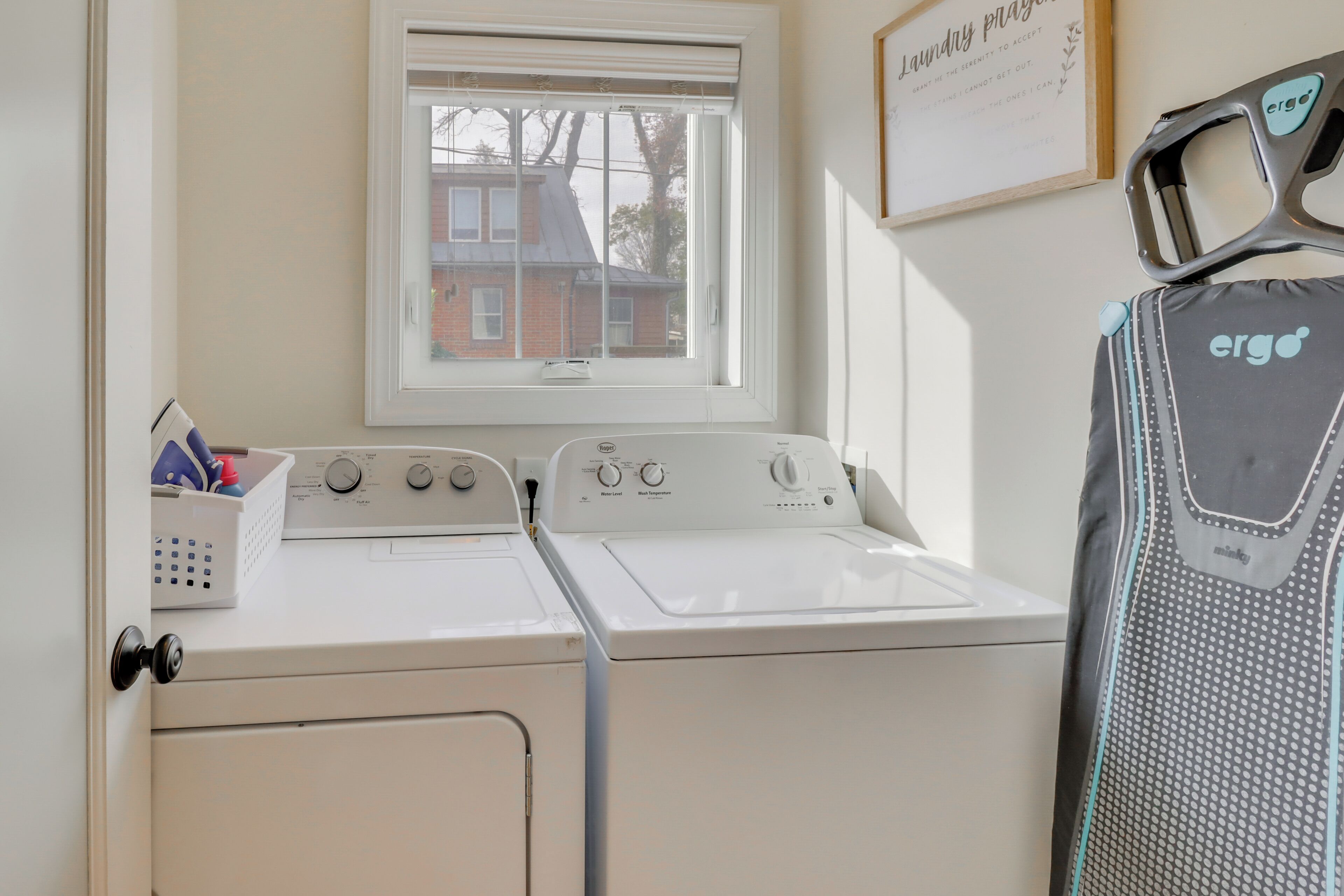 A laundry room with a washer and dryer, a window with a view of a house, and a framed sign on the wall.