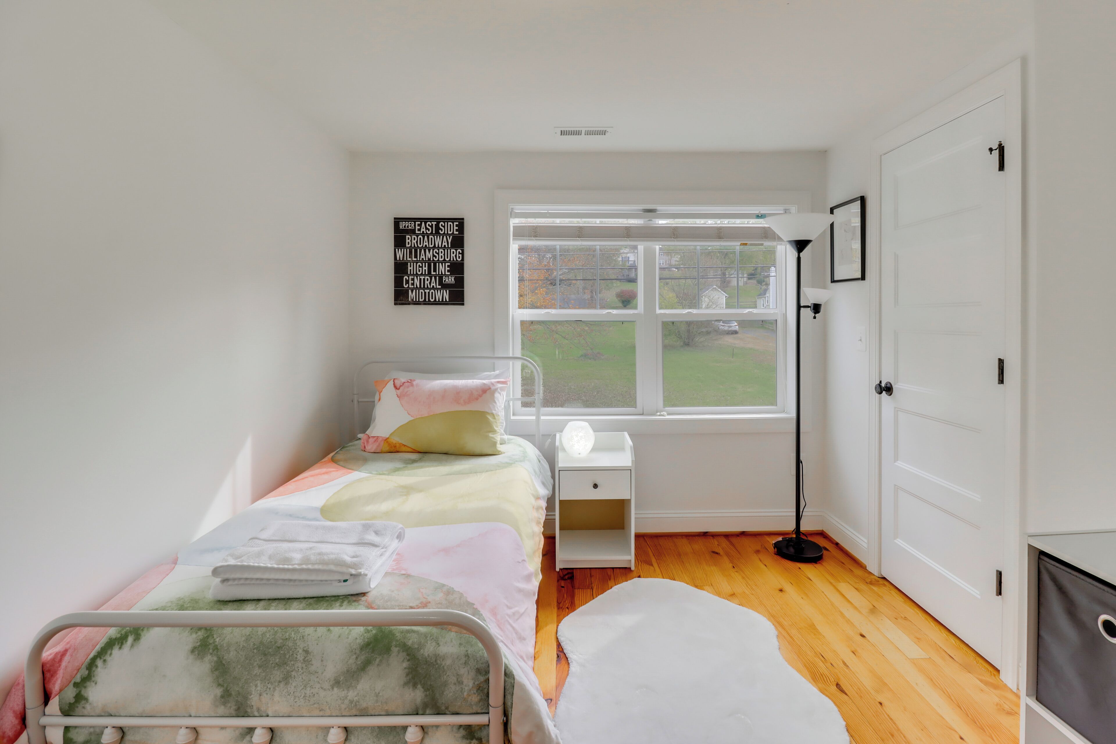 A bedroom with a bed, bedside table, lamp, and a window with a view of the outside.