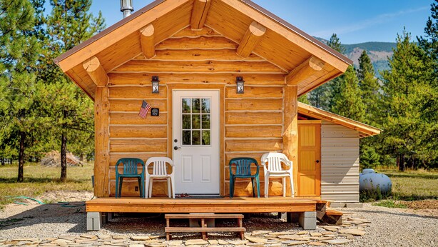 A log cabin with a front porch, two chairs, a bench, and a small shed. - Mtn Views & Fireplace: Cozy Trout Creek Tiny Home (Trout Creek)