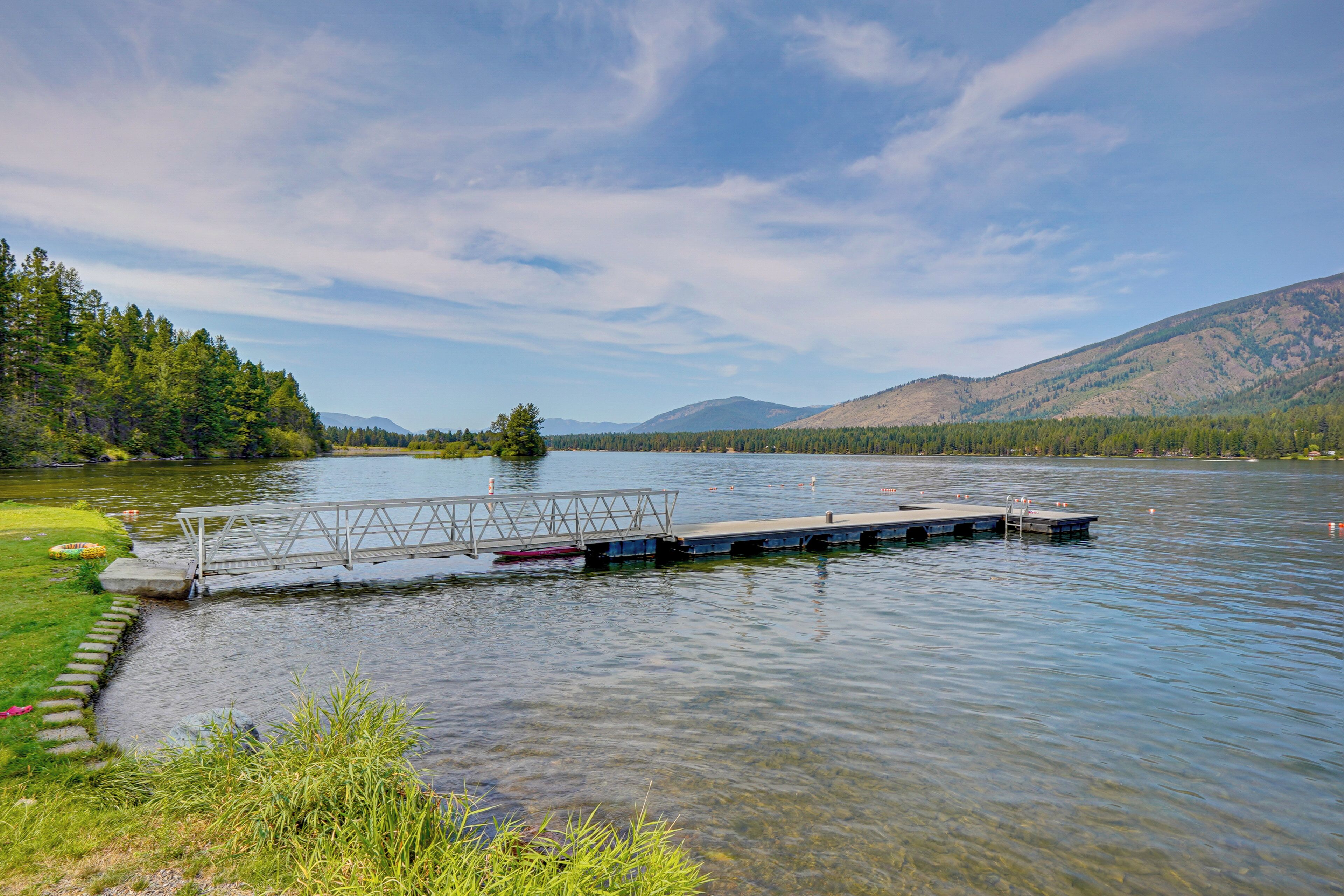 A calm lake with a wooden dock extending into the water, surrounded by lush greenery and distant mountains.