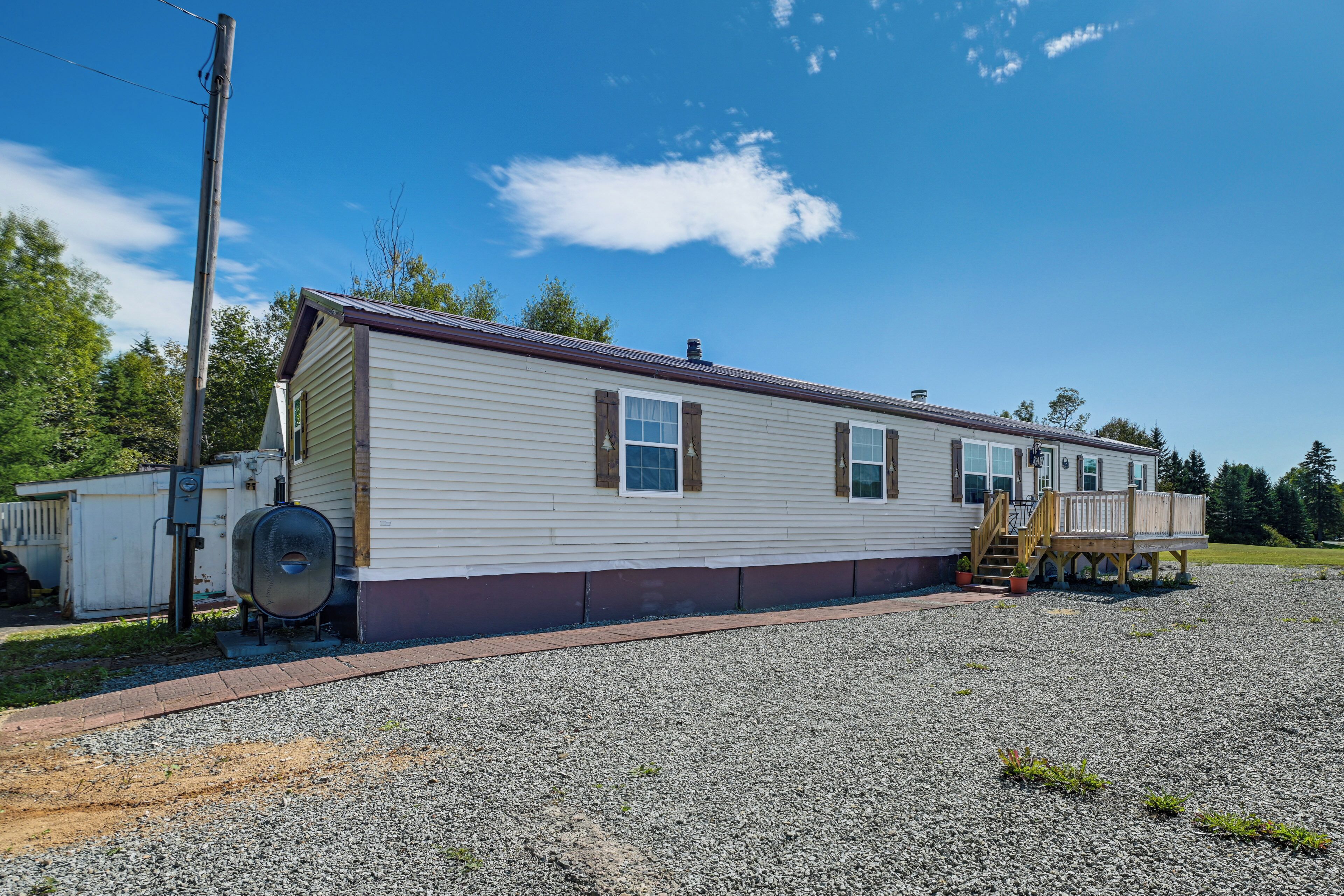A single-story mobile home with a gravel driveway, a wooden deck, and a utility pole.