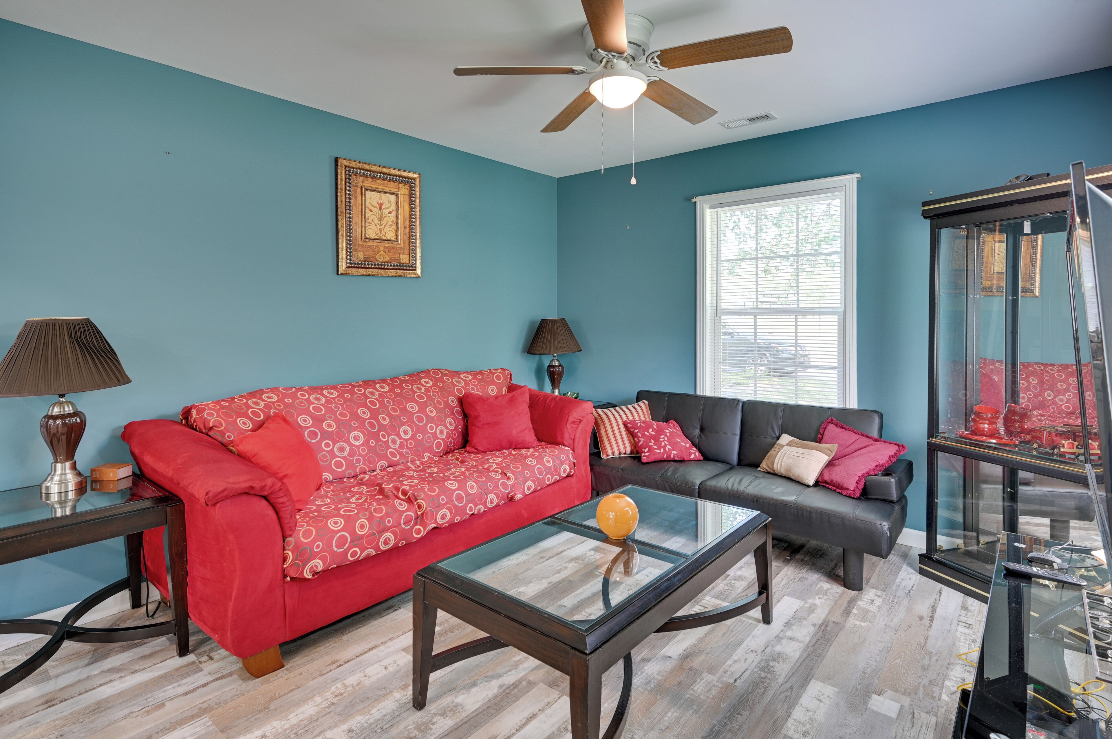 A living room with teal walls, a red patterned sofa, a black sofa, a glass coffee table, and a ceiling fan.