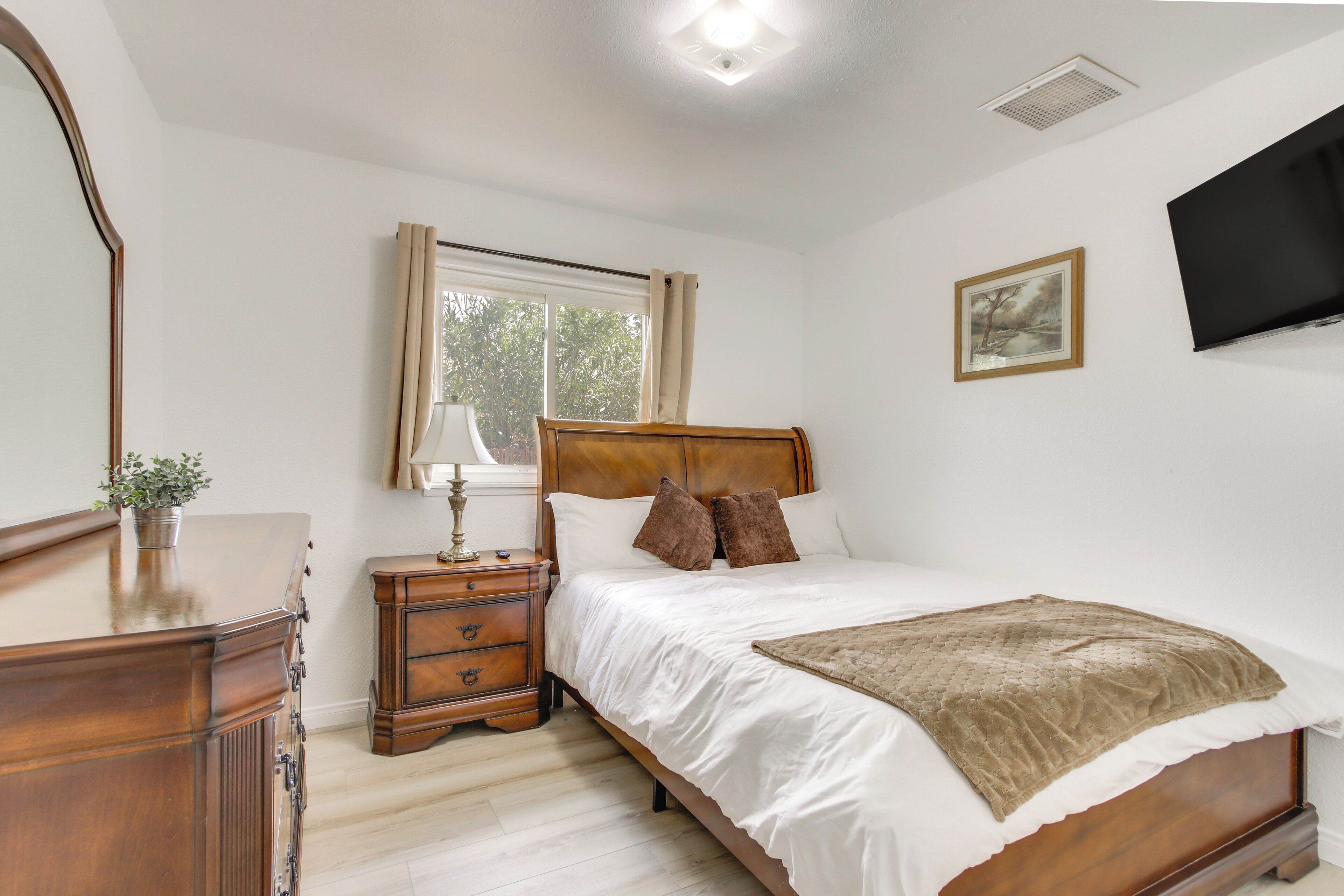 A bedroom with a wooden bed, a nightstand, a dresser, a window with curtains, a flat-screen TV, and a framed picture on the wall.