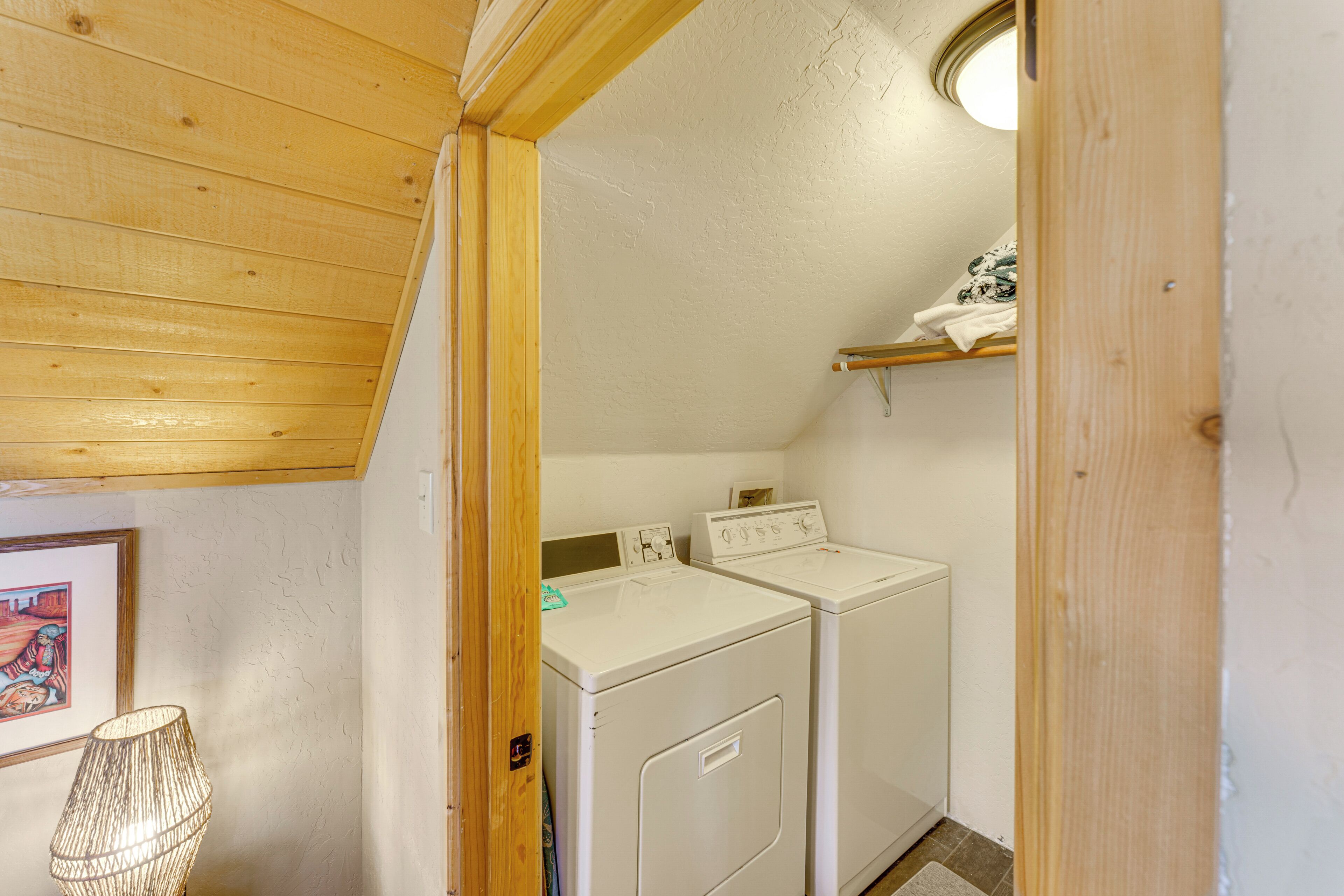 A laundry room with two white washing machines, a wooden ceiling, and a small shelf with folded clothes.