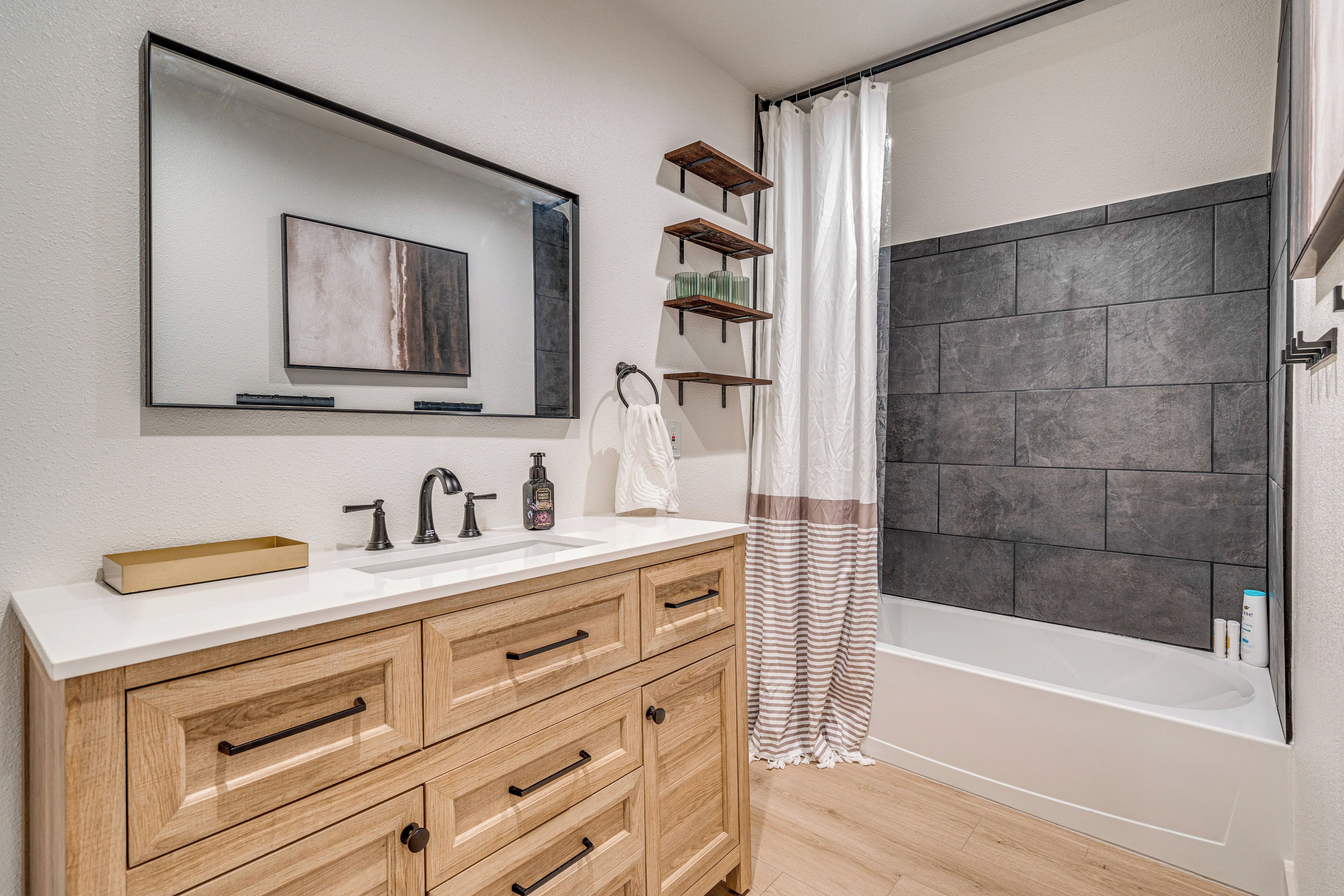 A modern bathroom with a white sink, wooden vanity, a large mirror, and a bathtub with a dark tiled wall.
