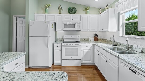 Una cocina moderna con armarios blancos, horno empotrado, microondas y lavavajillas. Hay jarrones decorativos en la pared y una ventana con cortinas.