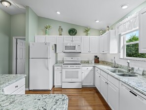 A modern kitchen with white cabinets, a built-in oven, microwave, and dishwasher. There are decorative vases on the wall and a window with curtains. - Stylish 3/2 Designer Home in Hemingway: Comfort, Luxury, and Perfect Location! (The Villages)