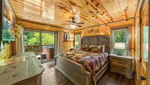 A wooden cabin bedroom with a bed, a nightstand, a dresser, a ceiling fan, and a window overlooking greenery.