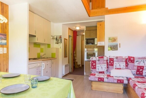 Une pièce confortable avec une table à manger dressée pour quatre personnes, une kitchenette équipée d’un four à micro-ondes et d’un four traditionnel, et un canapé à motifs.
