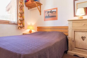 Un lit bien fait, recouvert d’une couvre-lit à motifs, une table de chevet en bois avec une lampe, un tableau accroché au mur et une armoire en bois ornée d’une découpe en forme de cœur.