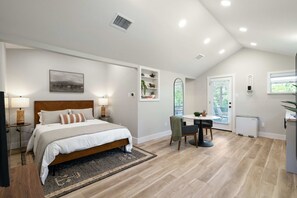 A bedroom with a large bed, bedside tables, a desk, and a chair. There is a window with blinds, a radiator, and a small plant on the desk.