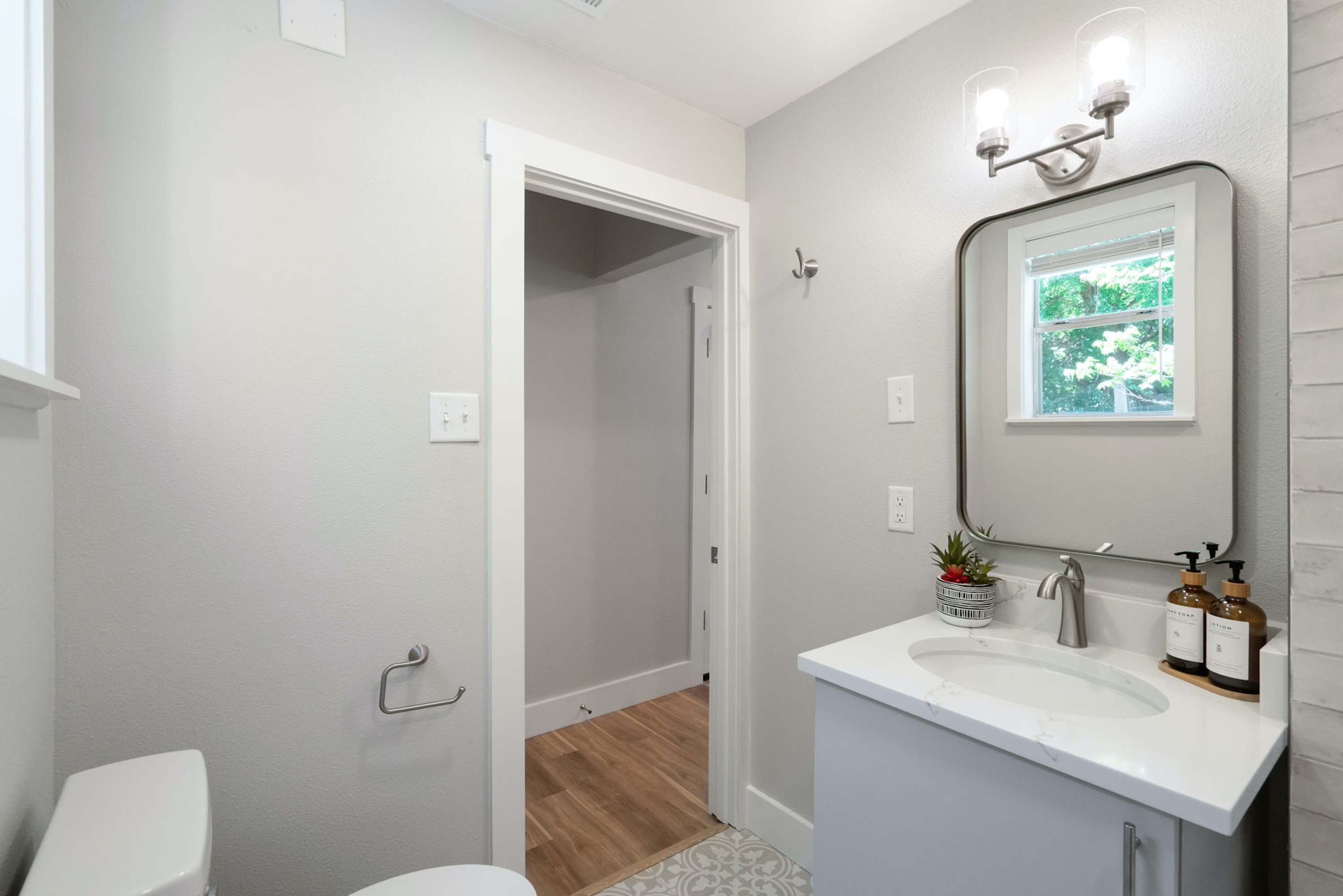 A bathroom with a white sink, mirror, and toilet. There is a small plant on the sink and a window above the mirror.