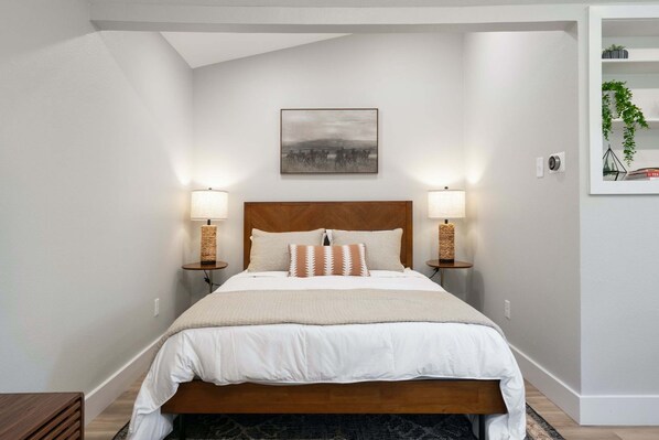 A neatly arranged bedroom with a wooden headboard, two bedside lamps, a large bed with white bedding, and a framed picture on the wall.