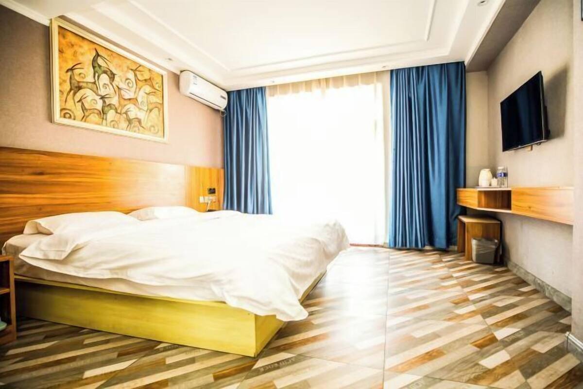 A hotel room with a large bed, a wooden headboard, a flat-screen TV, a painting on the wall, and a window with blue curtains.