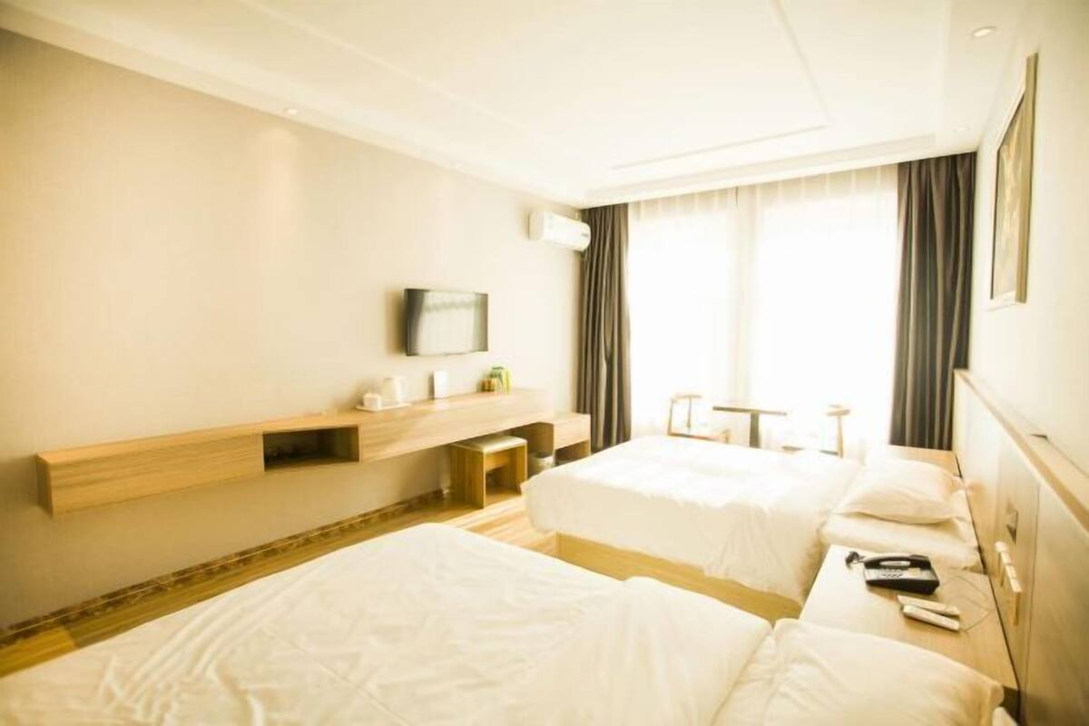 A hotel room with two beds, a wooden bedside table, a mounted TV, and a window with curtains.