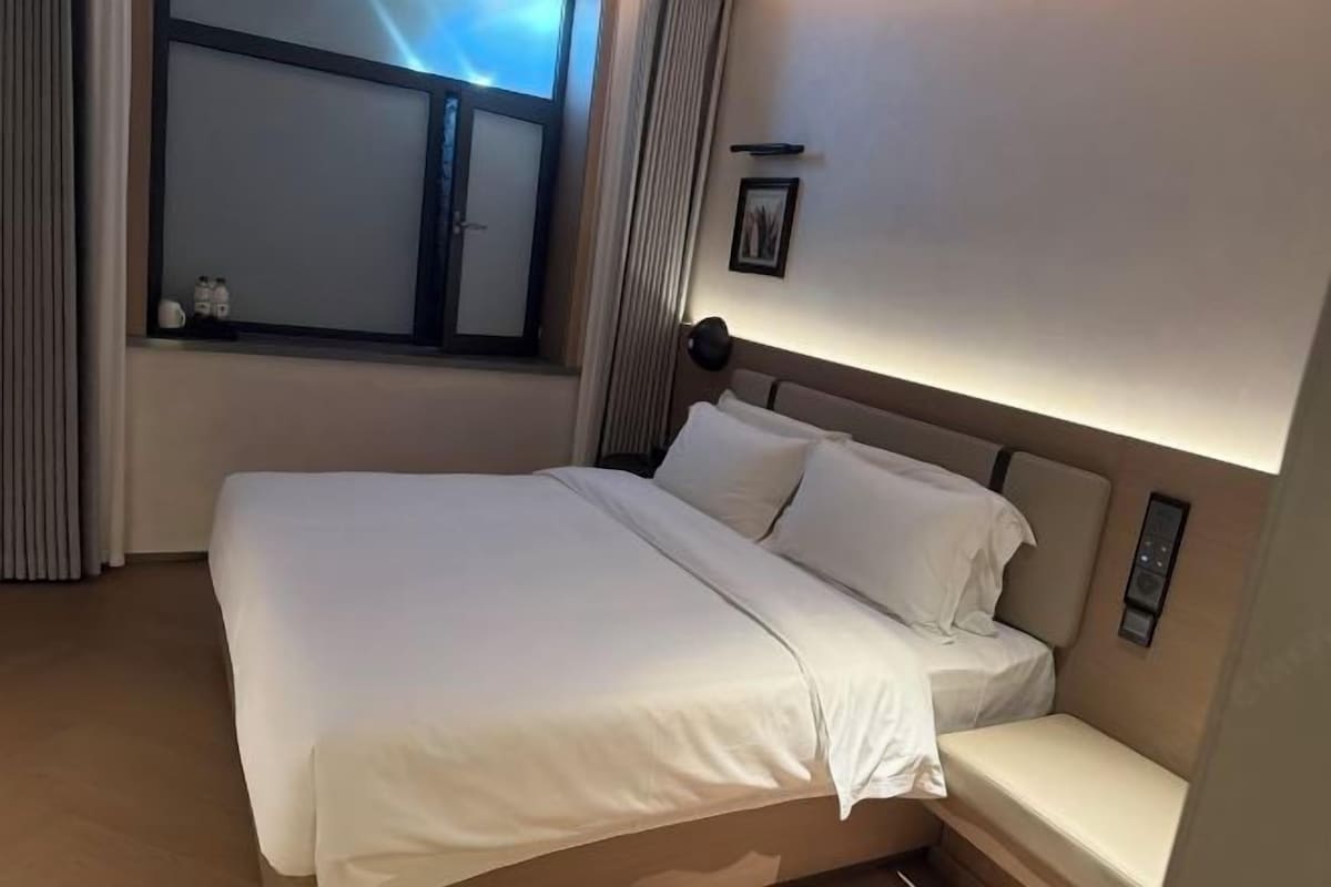 A hotel room with a bed, a window with curtains, a bedside table, and a wall-mounted control panel.