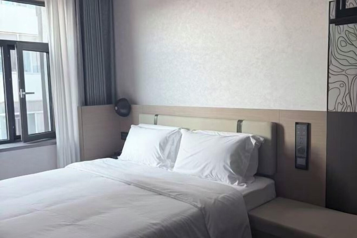 A hotel room with a bed, a bedside table, a window with curtains, and a wall-mounted control panel.