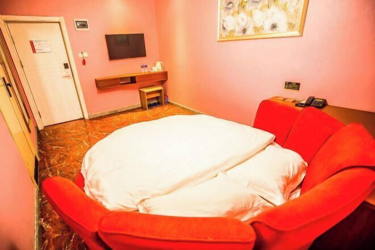 A hotel room with a bed, a red armchair, a small table with a telephone, a TV on the wall, and a painting.