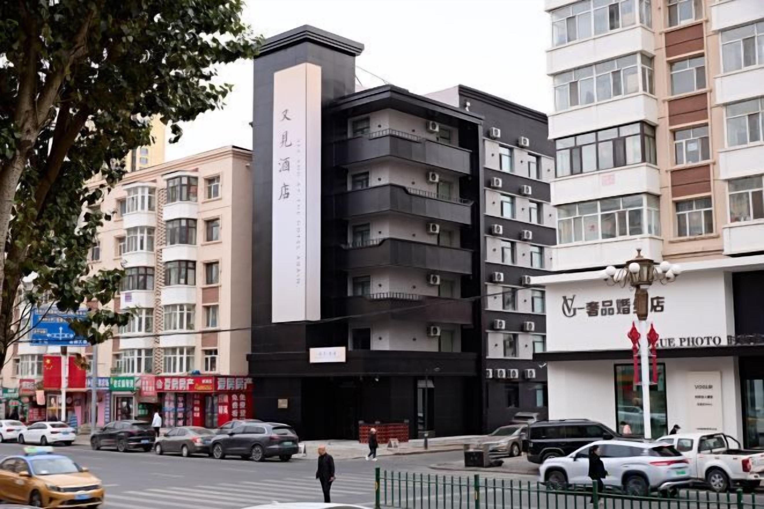 Un edificio de varios pisos con un letrero vertical llamativo, un escaparate con toldo rojo y coches aparcados en la calle.