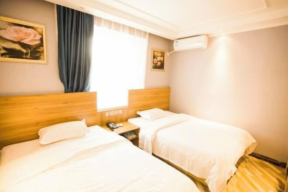 A hotel room with two beds, a wooden headboard, a window with curtains, an air conditioning unit, and a small bedside table with a phone and shoes.