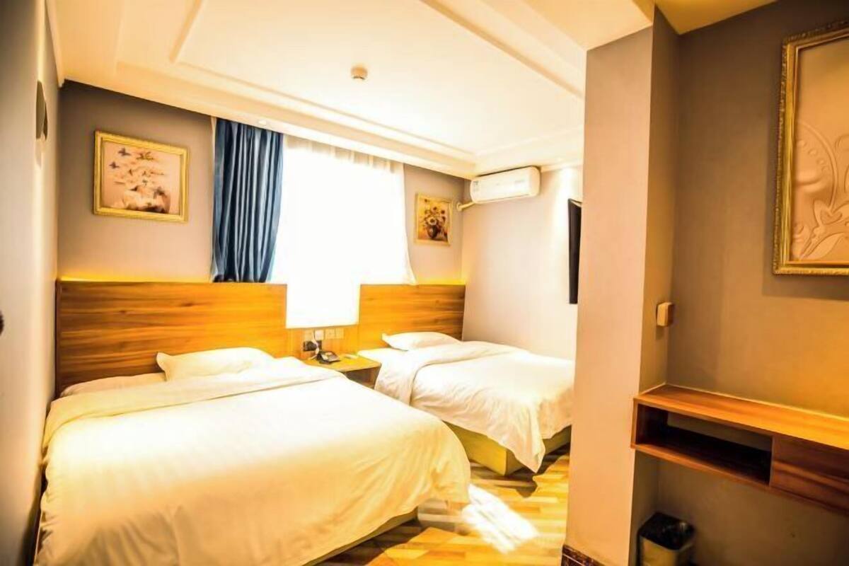 A hotel room with two beds, a wooden headboard, a window with curtains, and a wall-mounted air conditioner.