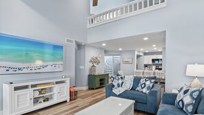 Un salon spacieux agrémenté d’un grand tableau représentant une plage, d’une console blanche, d’un canapé bleu et d’un escalier menant à un niveau supérieur.