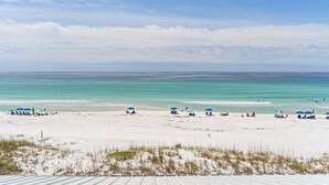 Une plage de sable blanc bordée d’une eau turquoise et limpide. On y trouve plusieurs parasols bleus et des chaises de plage.