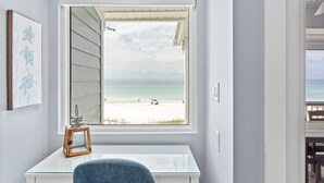 Une pièce dotée d’une grande fenêtre donnant sur une plage, d’un bureau et d’une chaise.