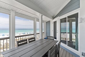 Un espace extérieur abrité, doté d’une table et de chaises, qui offre une vue sur une plage parsemée de parasols et fréquentée par des gens.