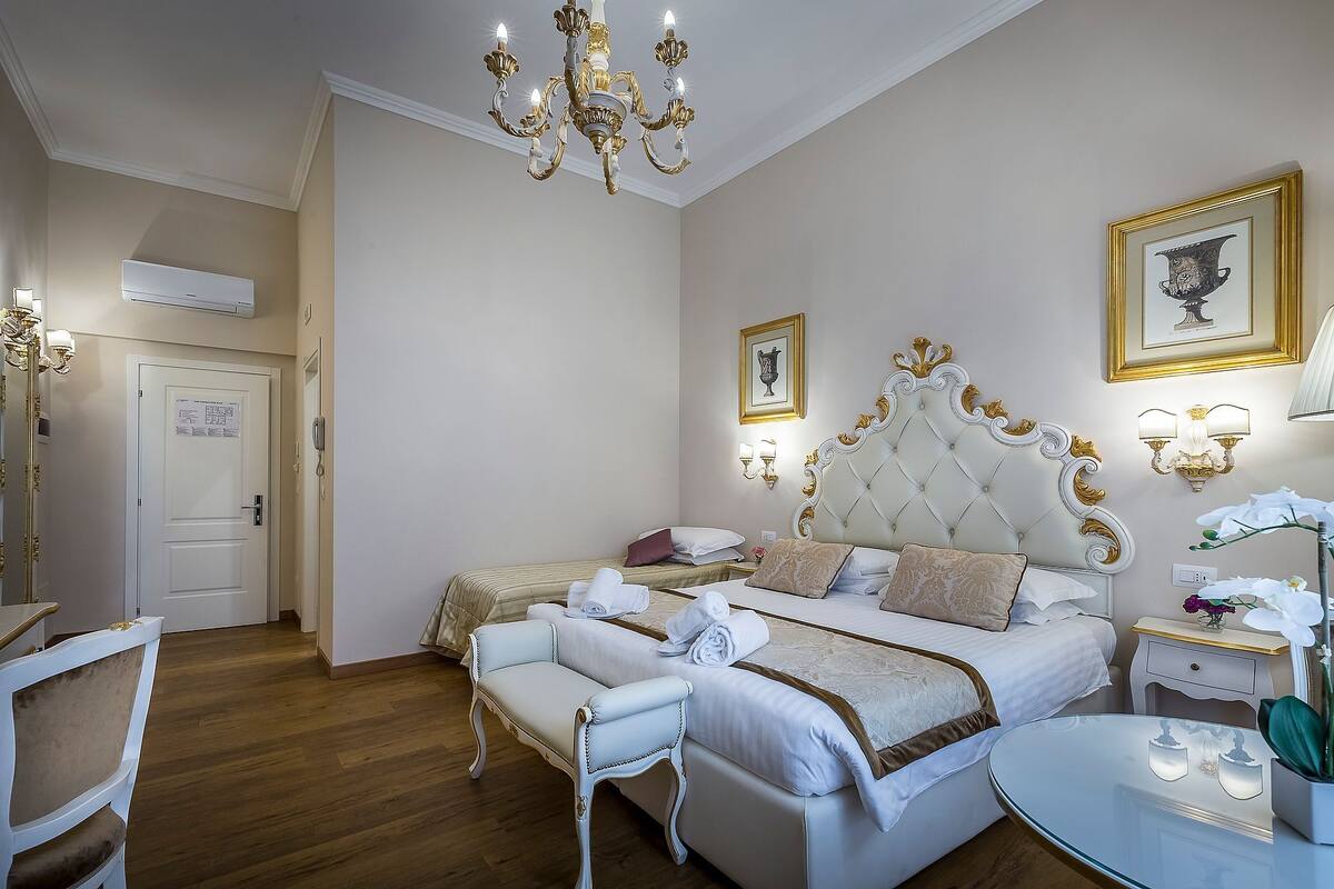A hotel room with a large bed, a sitting area with a chair, a glass table, and a chandelier.