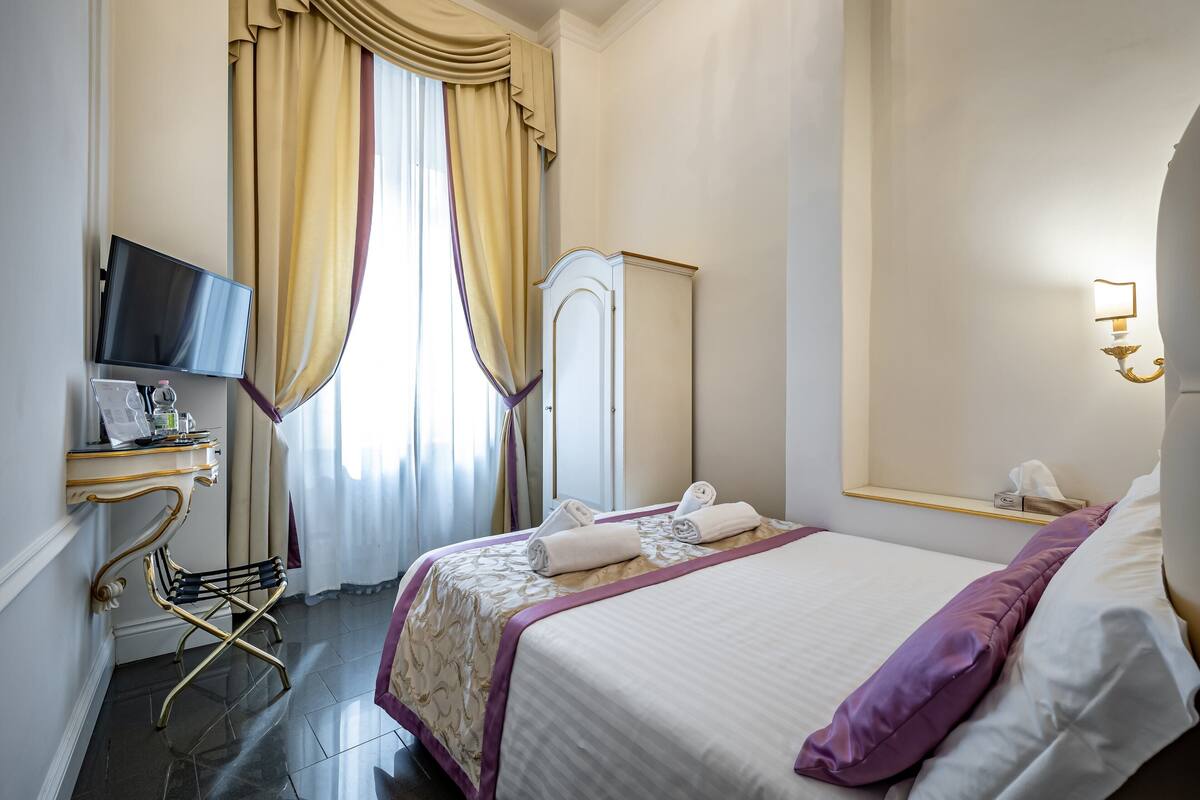 A hotel room with a bed, a desk with a computer, a wardrobe, and a window with curtains.