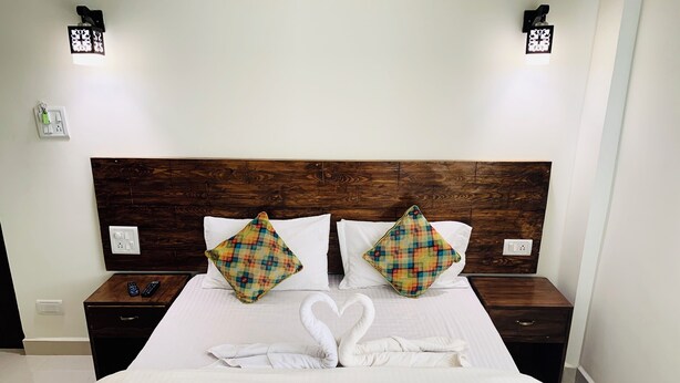 A neatly made bed with a white comforter and two pillows, a wooden headboard, bedside tables with remote controls, and wall-mounted light fixtures. - Mamata Courtyard Anjuna (Anjuna)