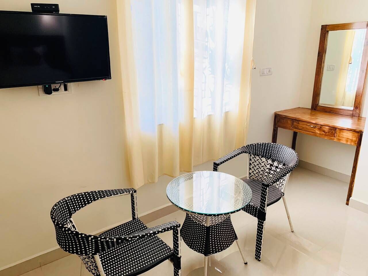A room with a flat-screen TV mounted on the wall, a round glass table with four chairs, and a wooden desk with a mirror.