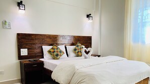 A neatly made bed with white linens, a wooden headboard, and two decorative pillows. There are bedside tables with remote controls and a wall socket. - Mamata Courtyard Anjuna (Anjuna)